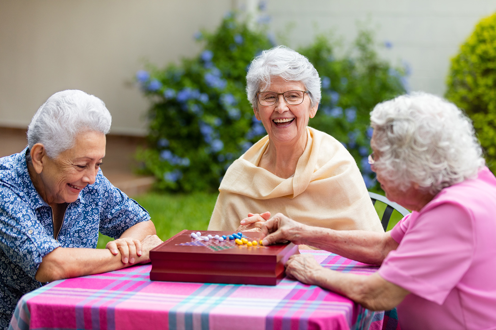 Cheerful latin elderly women sitting at a table outside, playing a boardgame and smiling at each other.