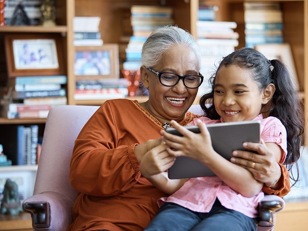 Smiling grandmother and granddaughter using digital tablet. Females are wearing casuals. They are sitting in living room at home.