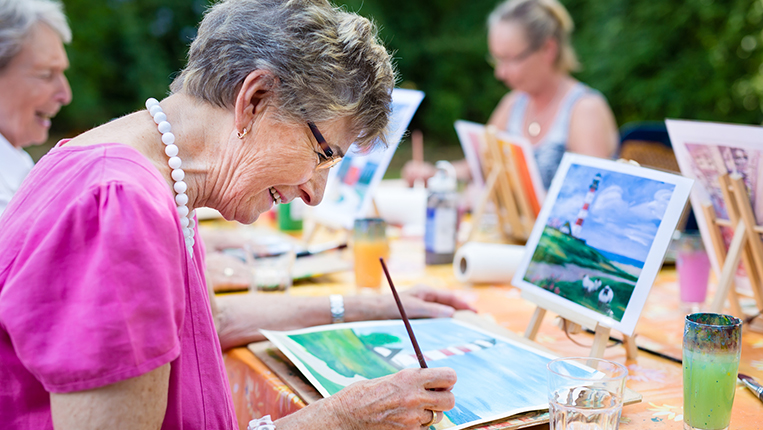 Side view of a happy senior woman smiling while drawing as a recreational activity or therapy outdoors together with the group of retired women.