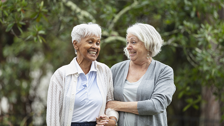 Two multiracial senior women conversing, walking together outdoors in a back yard, with trees behind them. The African-American woman is in her 70s. Her friend is in her 60s.