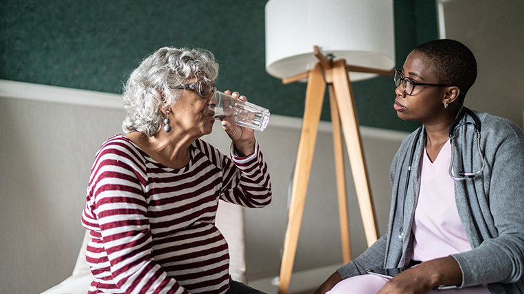 Senior woman drinking water with a nurse's help in the bedroom