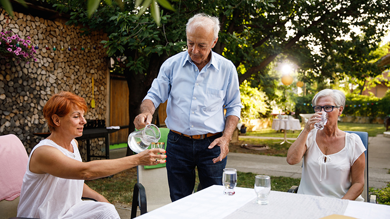 Candid shot of chivalrous senior man pouring a glass of water for his friend while hosting a dinner party in his back yard.