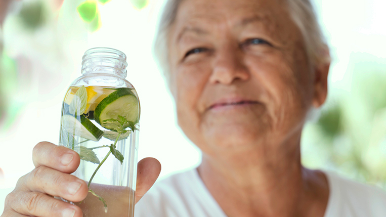 Senior female is holding a bottle of infused water with lemon slices, cucumber and rosemary leaf in it.