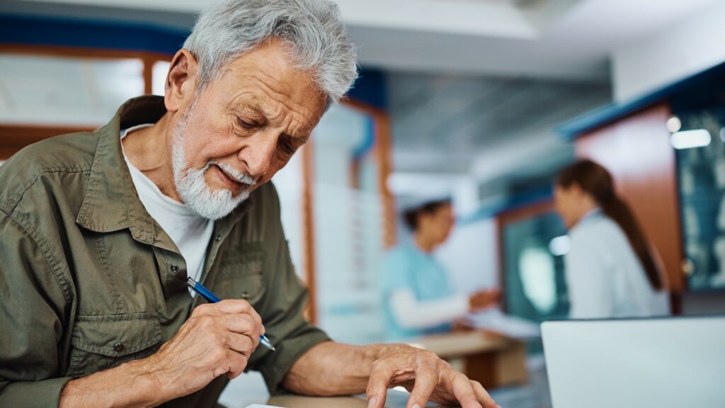 Senior man writing his data in medical report at reception desk in the hospital.