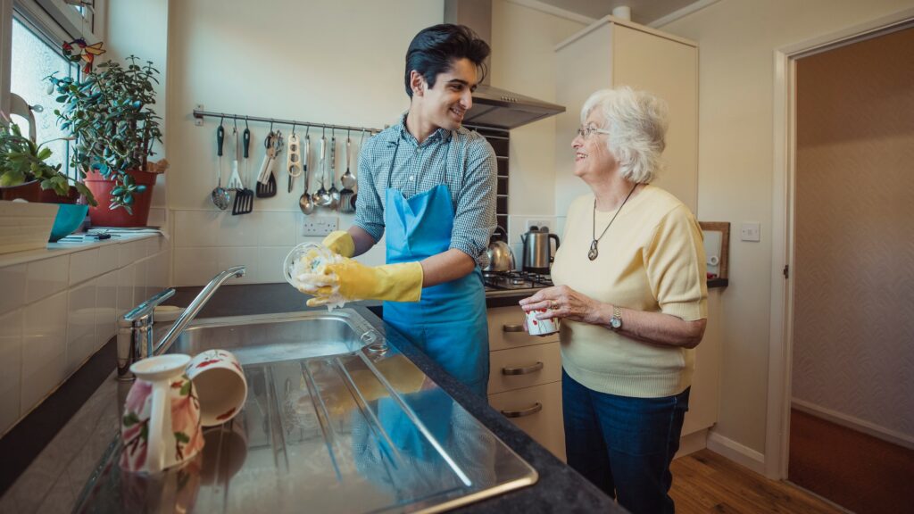 A home health aid washes dishes while talking to an older woman. 