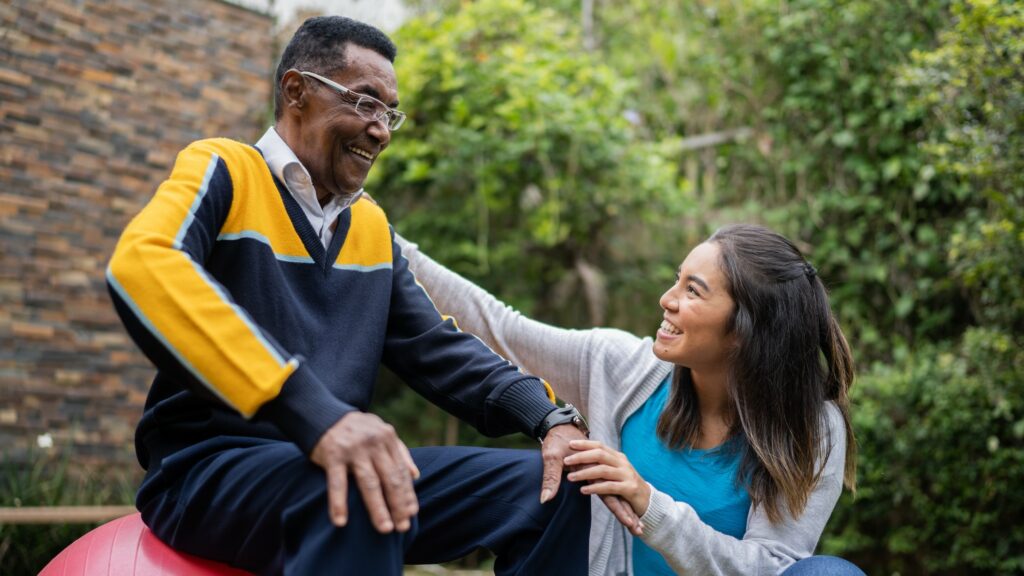 Physical therapist talking to senior man sitting on a fitness ball at home