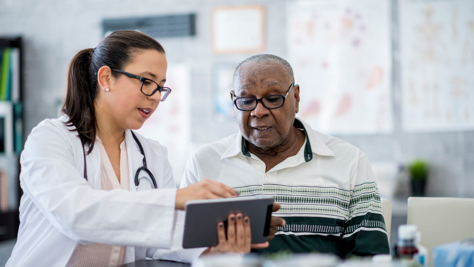 A senior man of African descent is indoors in a hospital room. He is watching his female doctor using a tablet computer. She is explaining a medication schedule to him.