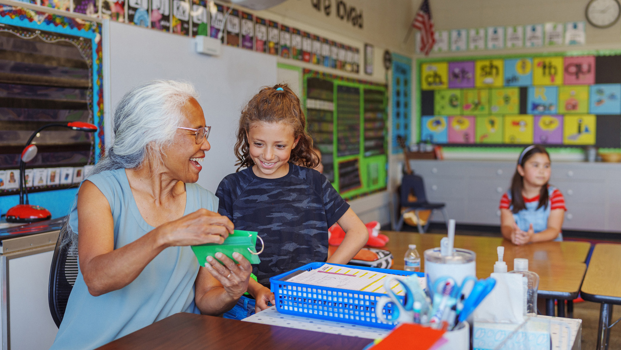 A cheerful and supportive mature adult female teacher of Pacific Islander descent sits at her desk in her classroom and provides one on one assistance to a smiling elementary age girl.