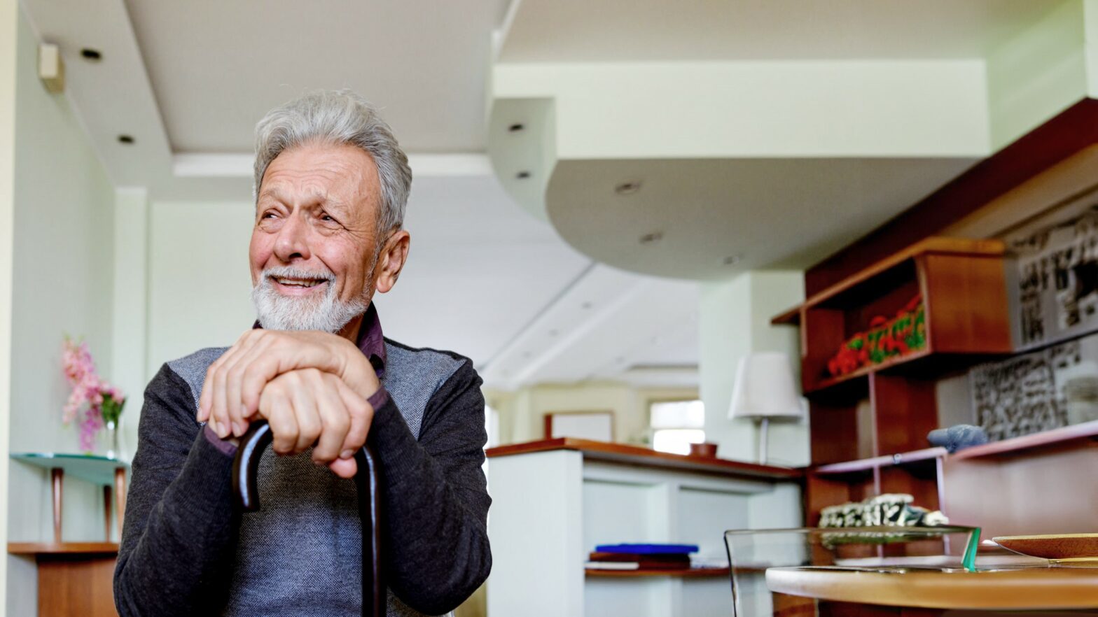 A senior Caucasian man is sitting at home, leaning against his cane and looking away with a warm smile on his face.