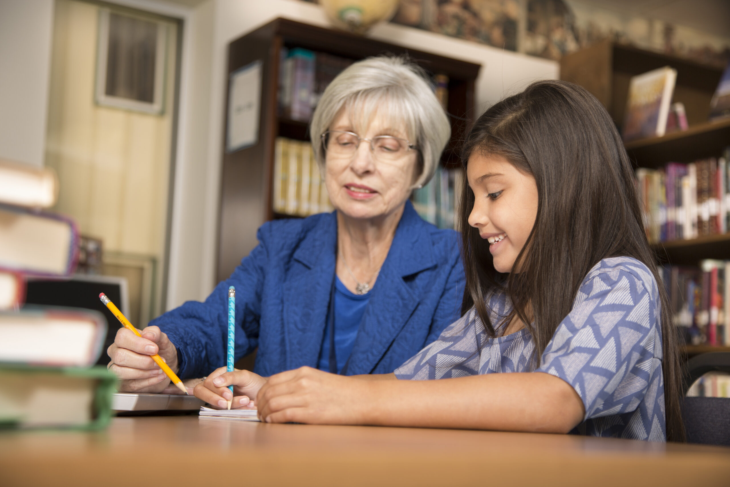 Senior adult teacher, mentor, or tutor assists Latin descent elementary age girl with homework in school classroom or library setting.